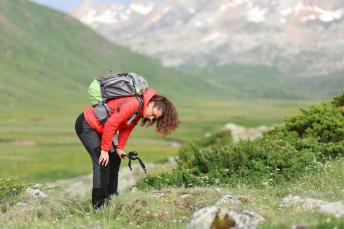 Turistka v červenej bunde s batohom odpočívajúca na horskej lúke počas celodenného trekingu.